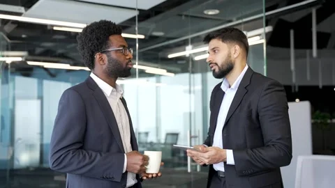 Two businessmen standing in modern office lobby holding coffee cups, discussing  Stock Footage 329052347