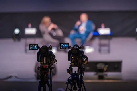 Two cameras capture live panel discussion with blurred speakers on stage and Stock Photos