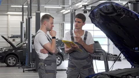 Two car mechanics engaged in a focused discussion over an open hood Stock Footage 311938545