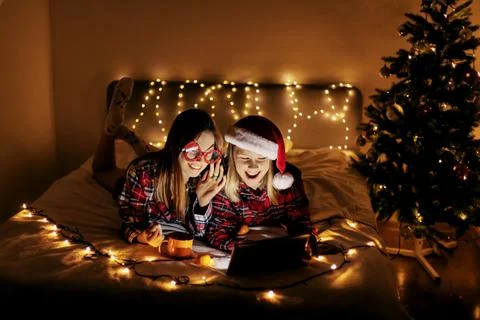 Two charming sisters lie on the bed with a laptop on Christmas or New Year's eve Stock Photos