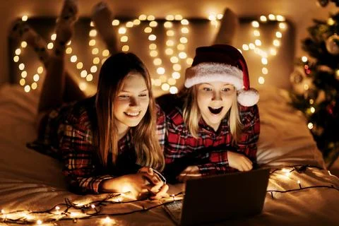 Two charming sisters lie on the bed with a laptop on Christmas or New Year's eve Stock Photos