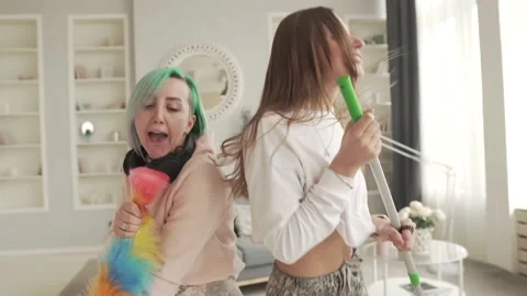 Two cheerful girls sing while cleaning the house. Friends have fun at home. Stock Footage 130601453