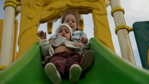 Two cheerful sisters slide down a green slide at a playground Stock Footage 306732977