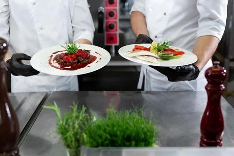 Two chefs in a professional kitchen hold ready-made dishes in their hands. Stock Photos
