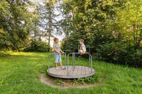 Two children are having fun on a merry-go-round in a lush park. They are smil Stock Photos