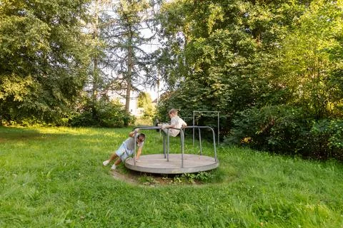 Two children are having fun outdoors, playing on a merry-go-round in a lush g Stock Photos