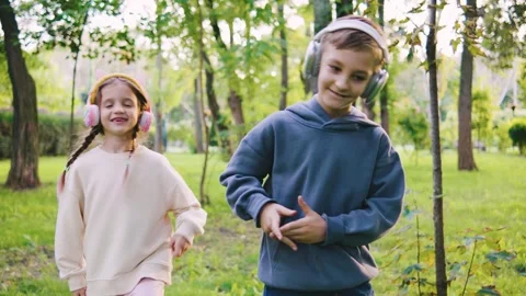 Two children dance joyfully in a park, wearing headphones and enjoying their Stock Footage 313751392