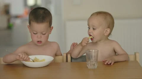 Two children having meal together, elder boy feeds toddler baby pasta from plate Stock Footage 219758074