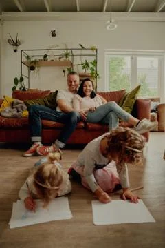 Two children lying on floor making drawing while parents enjoy watching them Stock Photos