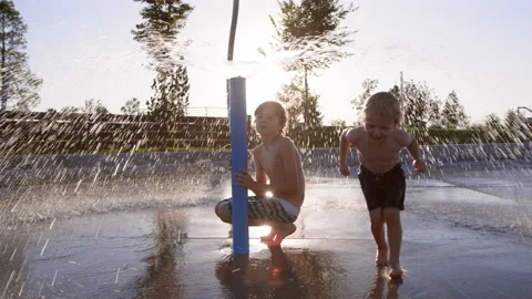 Two children play under large water sprinkler feature at splash pad, sunset 스톡 동영상 233940839