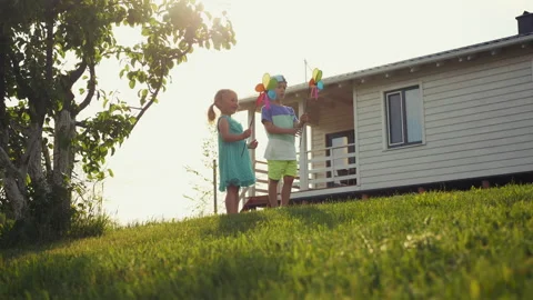 Two children play with windmills on a grassy slope near a house Stock Footage 277705550