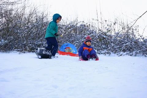 Two children playing with plastic sleds on snowy slope in winter Stock Photos