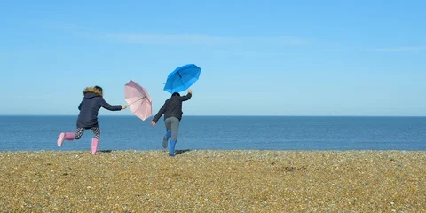 Two children playing with their umbrellas getting blown across a beach. Vídeo Stock 86424239