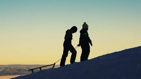 Two children pulling a sled running up a snowy hill during sunset Stock Footage 323390173