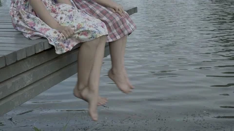 Two children shake their bare feet above the water. They sit on a wooden bridge  스톡 동영상 95447011
