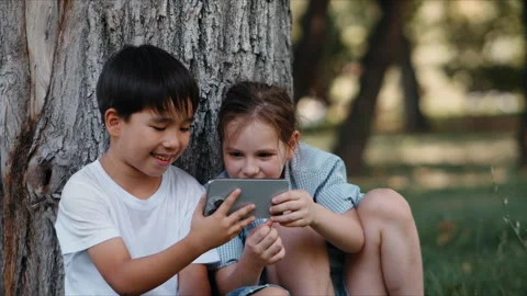 Two children smiling together as they play on a smartphone in a park near a tree Video stock 341879693