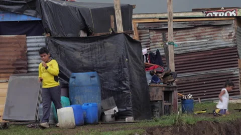 Two children standing outside an impoverished corregated metal and plastic home Stock Footage 146012332