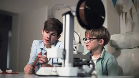 Two children at a table with a working 3D printer, discussing a printed part Stock Footage 335374295