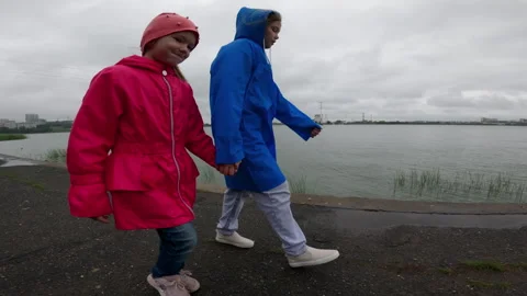 Two children in vibrant raincoats stroll along a tranquil lake on a cloudy day, Video stock 283673095