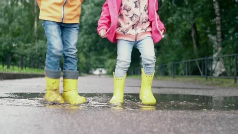 Two children in yellow rubber boots splash while standing in a puddle after the  Stock Footage