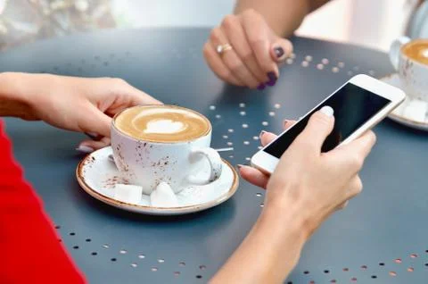 Two coffee cups and hands of young women with smartphone at outdoor cafe Stock Photos