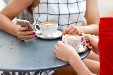 Two coffee cups and hands of young women with smartphone at outdoor cafe Stock Photos