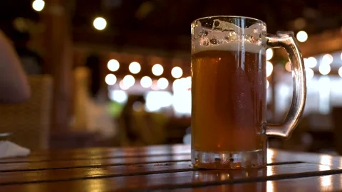 Two cold beer mugs on wooden table with blurred background lights and movemen Stock Footage 333619587