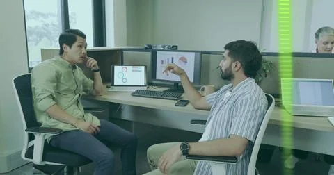 Two colleagues gesturing and discussing charts at office workstation with dual Stock Photos