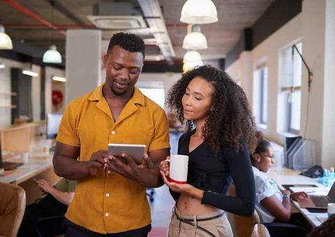 Two colleagues look at digital tablet in open plan diverse office Stock Photos