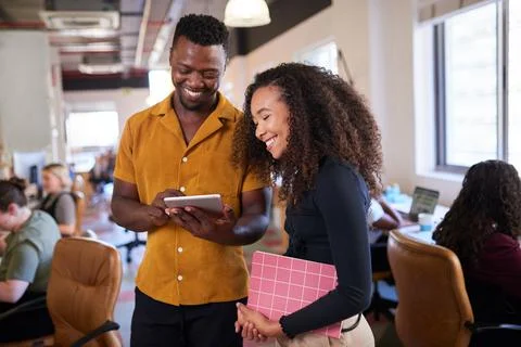 Two colleagues look at digital tablet in open plan diverse office Stock Photos
