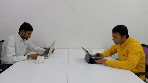 Two colleagues sit in front each other working on their computers, chatting and  Stock Footage 100025676