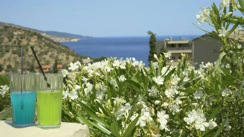 Two colorful drinks in front of white flowers with a coastal view, Greece Stock Footage 328675905