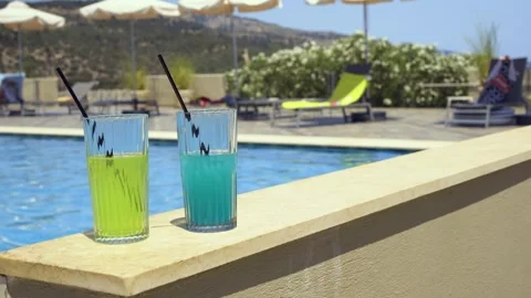 Two colorful drinks on a poolside ledge with loungers and umbrellas, Greece Stock Footage 328670834