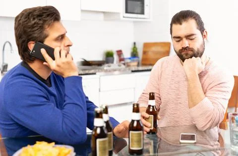 Two concerned men talking and laughing while enjoying beer at home, using phone Stockfoto's