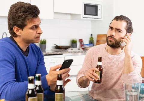 Two concerned men talking and laughing while enjoying beer at home, using phone Photos