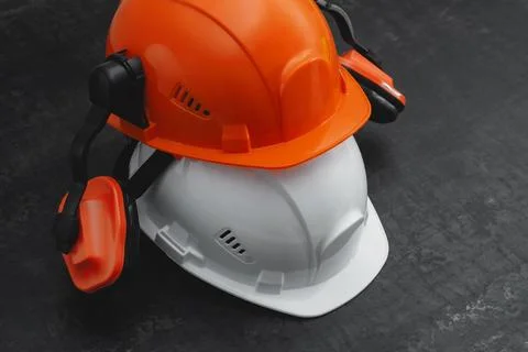 Two construction helmets, orange and white with earmuffs on a dark textured t Stock Photos