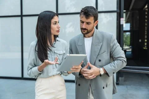 Two coworkers using a digital tablet and talking while standing outside. Mo.. Stock Photos