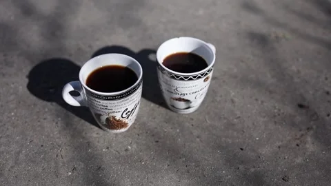 Two cups of black coffee rest on the rough pavement, bathed in sunlight and shad Stock Footage 304623211