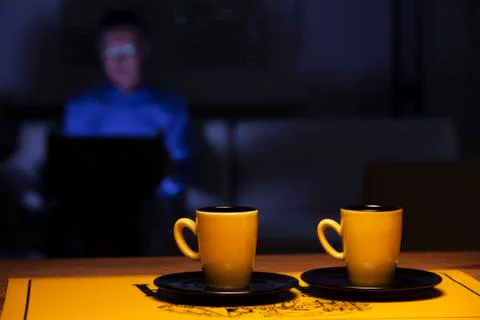 Two cups of coffee on a table and man with laptop on a couch in the backgroun Фото