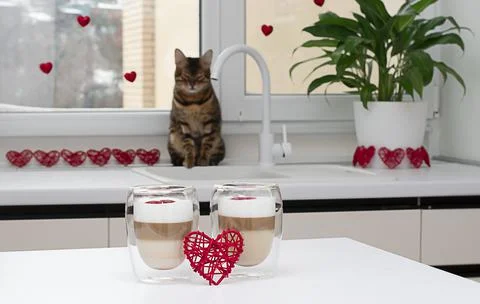 Two cups of latte on a white table against a window with red hearts. Stock Photos