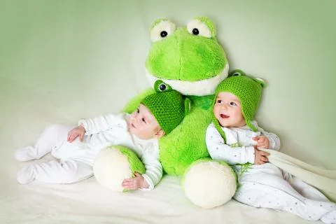 Two cute babies lying in frog hats with a soft toy Stock Photos