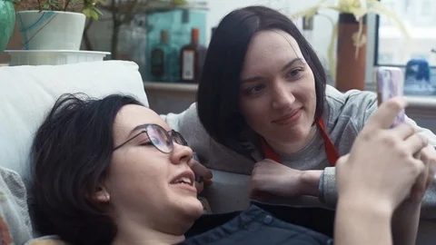 Two cute girls laughing using smartphone lying on couch at home. Stock Footage 87065563