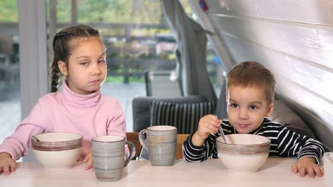 Two cute siblings sitting at dining table in country house kitchen eating Stock Footage 220674724