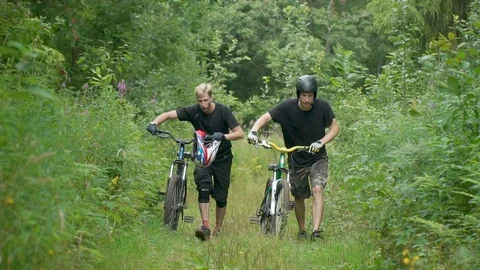 Two cyclists go uphill, carry bicycles nearby, all around grass trees and bushes Stock Footage 116488448