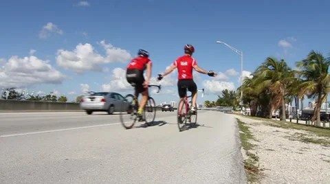 Two cyclists pass by on highway with palm trees in B/G Stock Footage 45522181