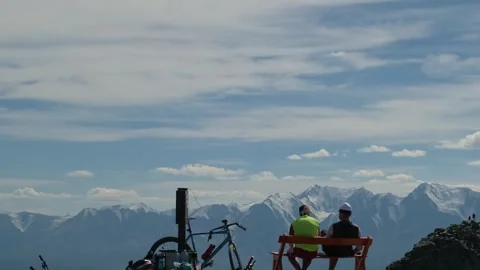 Two cyclists sitting on a mountain top with a stunning view of snowy peaks. Stock Footage 291772354