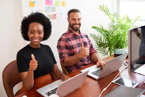 Two developers sitting at shared desk give thumbs up, coding in office on laptop 库存照片