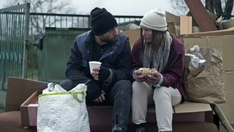 Two dirty and poorly dressed homeless people, a man and a woman, sit by a pile Stock Footage 226381116