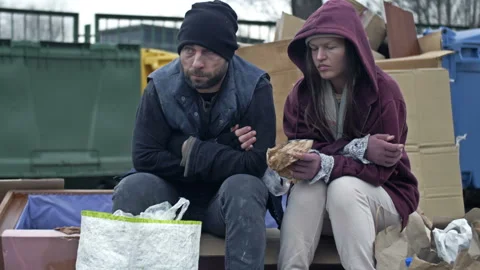 Two dirty and poorly dressed homeless people, a man and a woman, sit by a pile Stock Footage 230982215