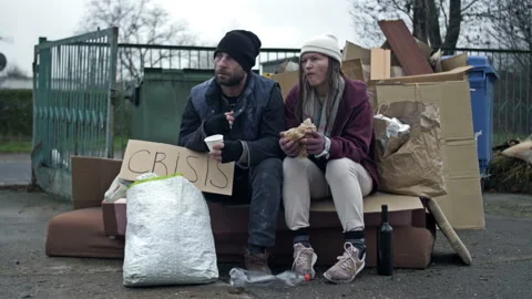 Two dirty and poorly dressed homeless people, a man and a woman, sit by a pile Stock Footage 240490590